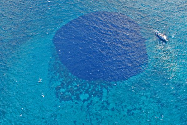 Photo courtesy of Chris Maio.  The research vessel Alucia sits near a blue hole in this image captured by a drone. The dark blue color signifies deep water surrounded by reefy shallows.