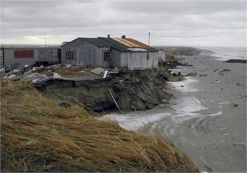 Photo by Tony Weyiouanna.  A house in Shishmaref, a village on Alaska's northwest coast, hangs over the edge of a cliff after Typhoon Tokage in 2004. The house fell onto the beach after erosion cut under it in subsequent years.