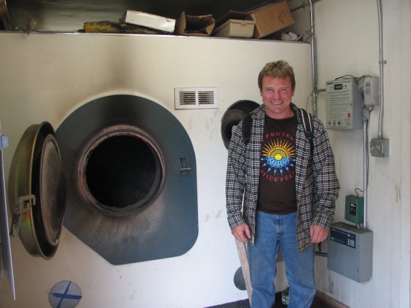 Photo by Karen Petersen. Jonathan Fitzpatrick of the Southeast Island School District poses with a biomass boiler in Thorne Bay that heats the school and its greenhouse and provides hot water.