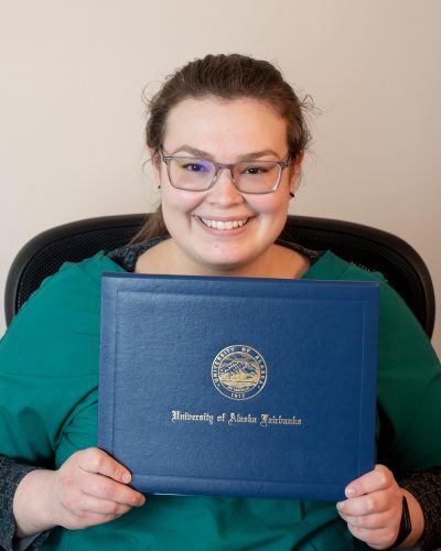 A young woman wearing glasses holds a UAF diploma cover.